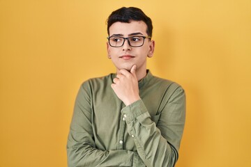 Non binary person standing over yellow background looking confident at the camera with smile with crossed arms and hand raised on chin. thinking positive.