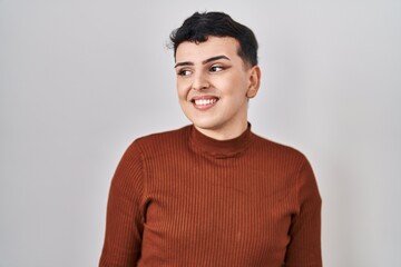 Non binary person wearing make up standing over isolated background looking away to side with smile on face, natural expression. laughing confident.