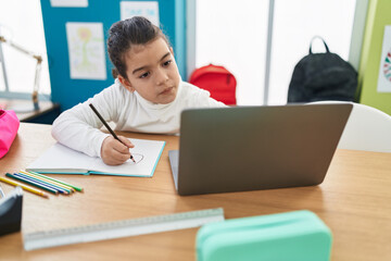 Adorable hispanic girl student writing on notebook using laptop at classroom