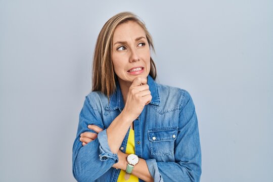 Young blonde woman standing over blue background thinking worried about a question, concerned and nervous with hand on chin