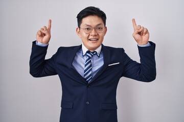 Young asian man wearing business suit and tie smiling amazed and surprised and pointing up with fingers and raised arms.