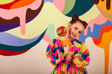 Fototapeta na wymiar Classy pretty little girl holds round lollipop at multi colored wall background, smiles looking at camera. Lovely child in colorful dress with candy on stick. Summer sweet concept. Copy ad text space