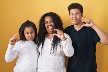 Family of mother, daughter and son standing over yellow background smiling and confident gesturing with hand doing small size sign with fingers looking and the camera. measure concept.