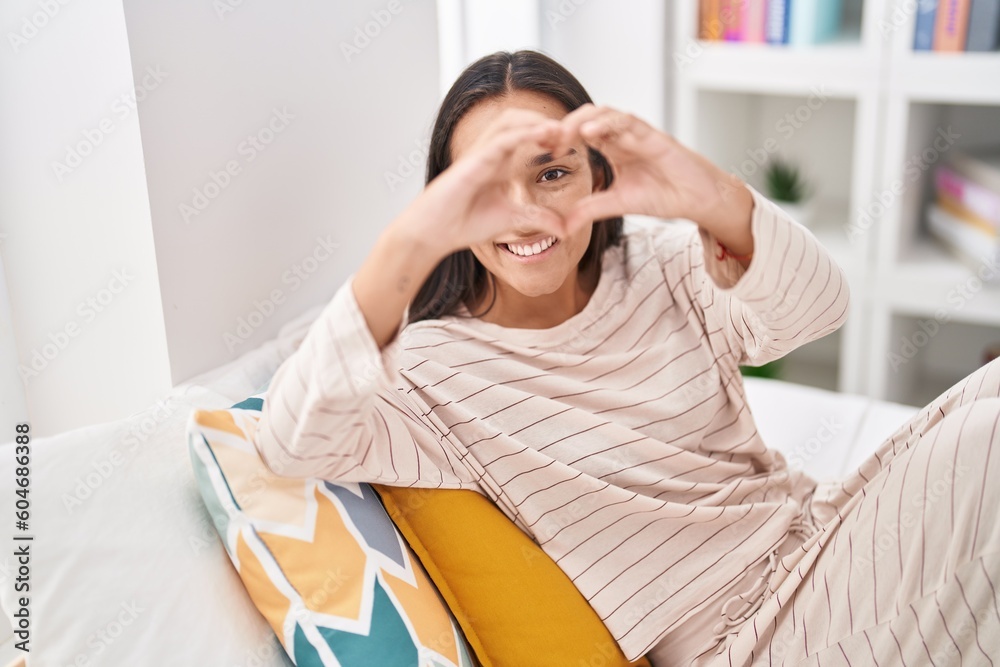 Sticker young beautiful hispanic woman doing heart gesture sitting on bed at bedroom