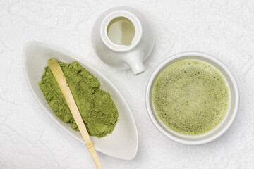 Matcha green tea in a bowl. Bamboo measuring spoon in a bowl of matcha powder. Porcelain milkman with milk.