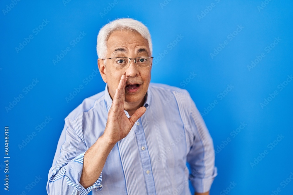 Poster Hispanic senior man wearing glasses hand on mouth telling secret rumor, whispering malicious talk conversation