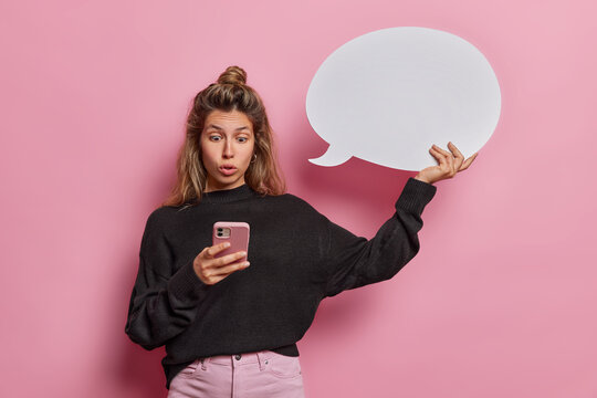 Stunned speechless young woman stares in smartphone with omg expression wears casual black jumper and trousers holds blank speech bubble isolated over pink background. Human reactions concept