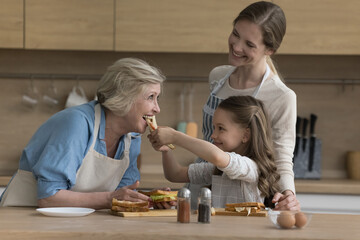 Preschooler cute girl feeds her granny with fresh prepared sandwich cooking together with young mother in cozy domestic kitchen. Homemade chores, communication and bond of multi-generational family