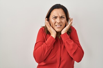 Hispanic mature woman standing over white background covering ears with fingers with annoyed...