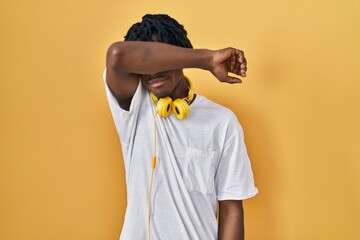 Young african man with dreadlocks standing over yellow background covering eyes with arm, looking...