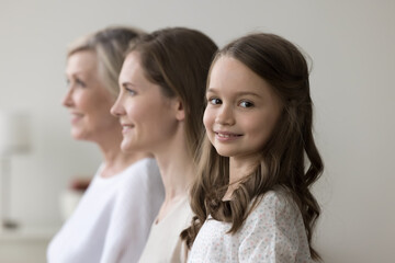 Happy preschooler girl staring at camera posing standing in row with young pretty mother and older grandmother look forward. Multigenerational family shooting indoor. Life from childhood to retirement