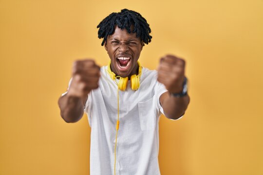 Young african man with dreadlocks standing over yellow background angry and mad raising fists frustrated and furious while shouting with anger. rage and aggressive concept.