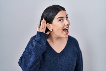 Young brunette woman standing over isolated background smiling with hand over ear listening an hearing to rumor or gossip. deafness concept.