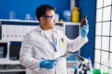 Young chinese man wearing scientist uniform reading clipboard at laboratory