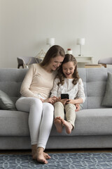 Little girl use mobile phone with young carefree mom sitting on couch at home in living room, looking at screen, smile enjoy new filter, spend leisure time together watch videos with parental control