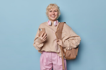 Fototapeta na wymiar Studio shot of cheerful blonde female college student carries backpack holds modern smartphone checks received notification uses headphones for listening news dressed casually isolated over blue wall
