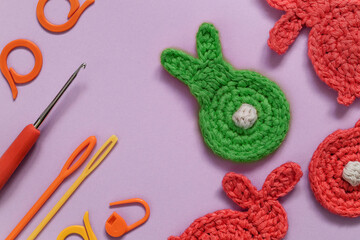 Crochet accessories and crochet green and red bunnies on a violet background. Top view. Copy space.