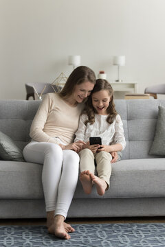 Little Girl Use Mobile Phone With Young Carefree Mom Sitting On Couch At Home In Living Room, Looking At Screen, Smile Enjoy New Filter, Spend Leisure Time Together Watch Videos With Parental Control