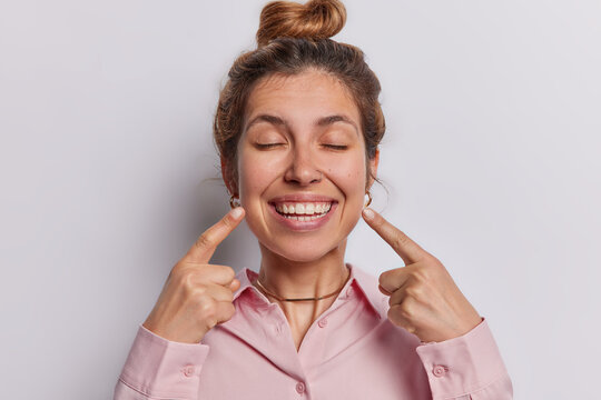 Photo of pleased adult woman points both index fingers at toothy smile has perfect even teeth keeps eyes closed dressed in pink shirt isolated over white background. Look at my perfect smile
