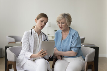 Focused female doctor in white coat showing to older woman patient health test results, explains prescription use medical app on digital tablet, give recommendation about disease treatment medications - obrazy, fototapety, plakaty