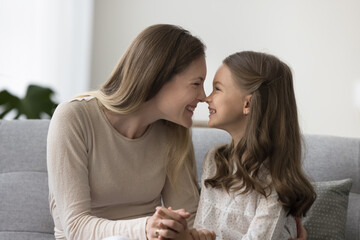 Loving young mom touch noses with cute daughter sit on sofa at home, enjoy funny moments, time together, hug with affection, laughing feel affection, two generation family feel love, happy motherhood