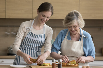 Millennial woman having pleasant weekend with mature mother, family wear aprons prepare handmade sandwiches for snack or breakfast, smile, enjoy pastime together and food preparation in cozy kitchen
