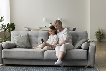 Cheerful grandmother and granddaughter sit on couch, use cellphone together, play on-line games,...