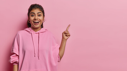 Happy excited Indian woman looks with great wonder at camera points index finger at upper right corner advertises something dressed in casual hoodie isolated over pink background. Hey check this out