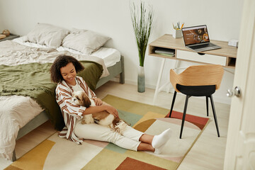 Full length portrait of happy young woman sitting on floor at home and cuddling with pet dog in...