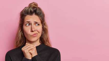 Embarrassed confused European woman keeps hands together purses lips doesnt know how to act wears casual black jumper concentrated aside poses against pink background copy space for your text - obrazy, fototapety, plakaty