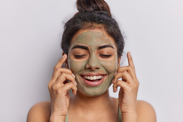 Cheerful brunette woman smiles broadly keeps eyes closed applies facial mask for hydrating skin stands bare shoulders isolated over white background pampers face. Beauty and wellness concept