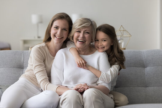 Three female generations family portrait. Cute little girl hugging tightly her beloved granny sit on sofa with young mom, smile look at camera, enjoy leisure time together. Unconditional love and ties