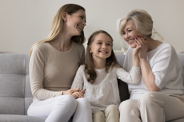 Happy multigenerational women family spend pastime, enjoy caress pleasant time together sit on sofa in living room. Older 60s, young 30s and younger 6s female relatives rest on weekend leisure at home