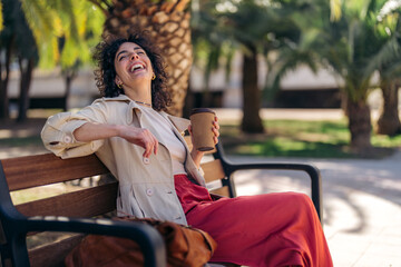 Smiling carefree young woman with curly hair sitting with legs crossed on bench in city park...