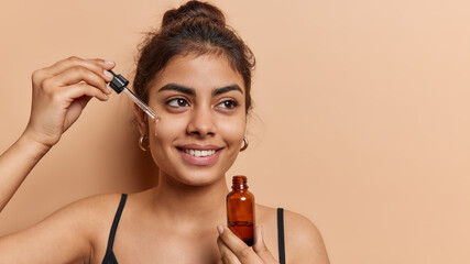 Domestic skin care and anti wrinkle routine. Pleased dark haired woman applies facial serum with dropper smiles toohthily focused aside dressed in t shirt isolated over brown background copy space - 604696943