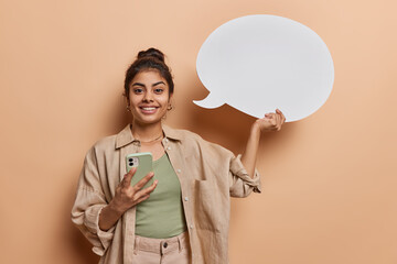 Positive good looking Indian woman smiles happily holds mobile phone and white speech bubble feels...