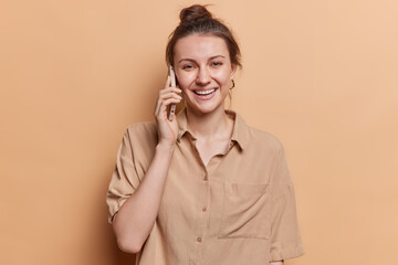 Communication concept. Cheerful young European woman with hair bun has phone conversation keeps smartphone near ear smiles positively dressed in casual shirt isolated over brown studio background.
