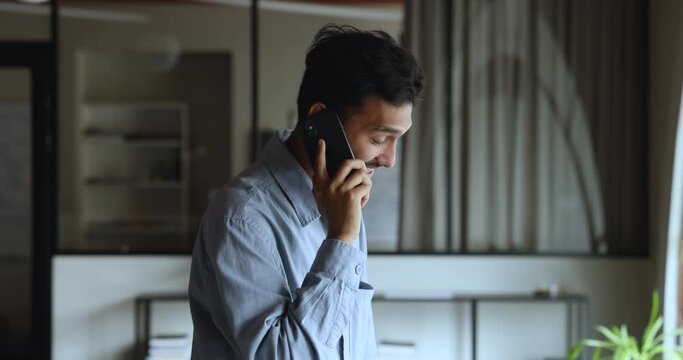 Successful Indian businessman lead conversation on cellphone stand in modern workplace, listen news, solves important issues with colleague, provide helpful information to client remotely. Phone call