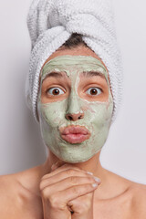 Vertical shot of surprised young woman has lips rounded hands under chin puts on beauty facial mask for skin cleansing pampering procedures stands topless after taking shower isolated on white wall