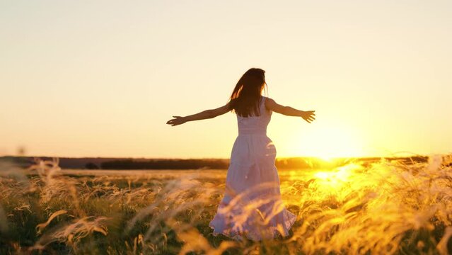 Young woman holding her arms to sides runs across field in sun. Concept of female dreams, success, travel, flight. Happy running girl. Free girl runs happily through meadow in grass in rays of sunset.