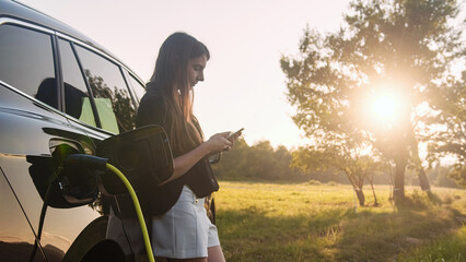Young woman browsing on a smartphone while waiting to charge an electric car outside the city in...