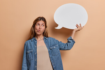 Thoughtful European woman with piercing in nose holds blank white speech bubble purses lips thinks what to write here dressed in denim shirt isolated over brown background. Let me think about it - obrazy, fototapety, plakaty