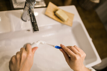 Fototapeta premium Female hand holding toothbrush with toothpaste applied on it in bathroom. Close up of female hand ready for brushing teeth. Young woman hand holding toothbrush with white tooth paste.