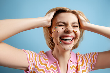 Excited woman with colorful rhinestones over her face holding hands in hair against blue background