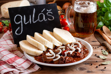 Traditional goulash meat with dumplings (knedliky) on white plate, tomatoes, onion, pepper, tablecloth in the background - typical Czech food