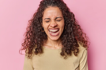 Photo of curly haired Brazilian woman keeps eyes closed exclaims loudly keeps mouth widwlt opened has white teeth dressed in casual brown jumper isolated over pink background. Human reactions concept