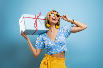Attractive young woman carrying a gift box on shoulder and smiling against blue background