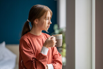 Teen moods. Upset teenage girl holding tea cup looking out window, feeling sad and anxious. Depressed worried teenager at home, unhappy adolescent thinking about life