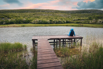 Hiker in blue jacket sitting on wooden pier by the lake with mountain in background at Burren national park, county Clare, Ireland, Adventure and lifestyle concept, wallpaper