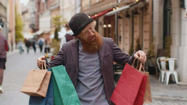 Happy smiling stylish beardedman shopaholic consumer after shopping sale with full bags with gifts outdoors. Redhead young adult guy tourist traveler walking in urban city street road. Town lifestyles